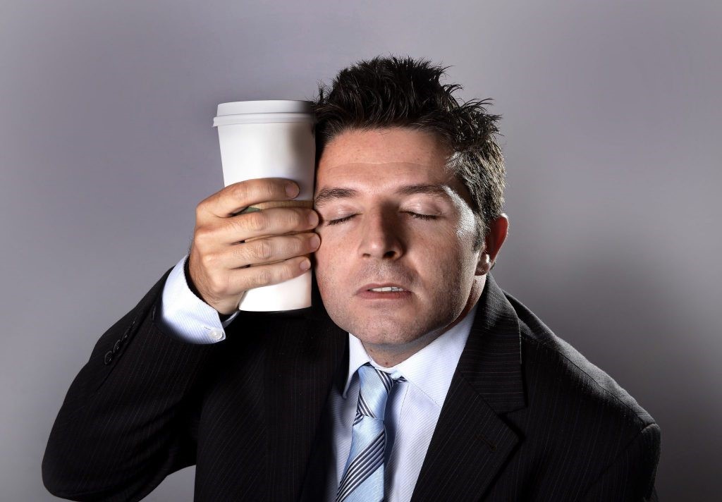 young sleepy addict business man in suit and tie holding cup of take away coffee against sleeping face in caffeine addiction and need to keep awake isolated on grey